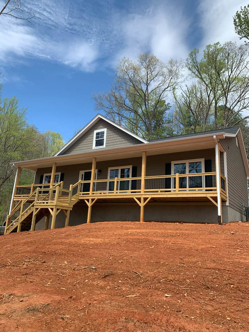 Wooden porch with railings and stairs overlooking a red dirt hill, surrounded by trees under a blue sky with scattered clouds