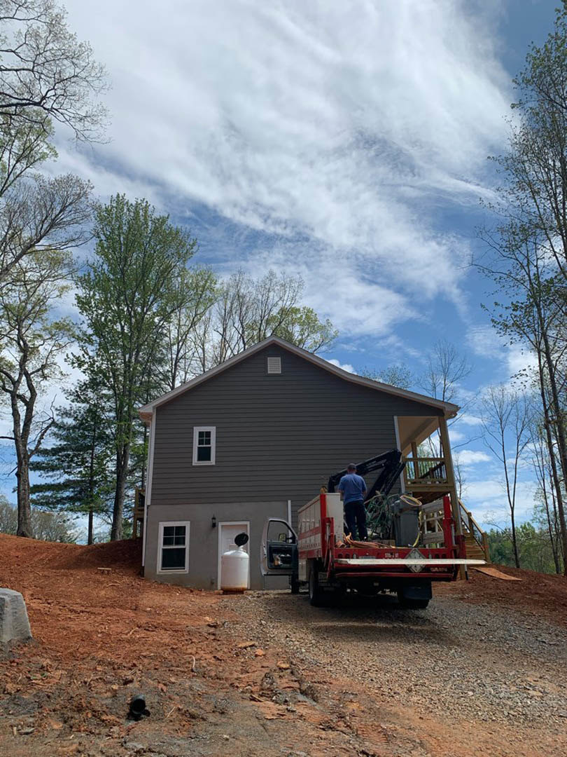 Dirt road leading to a custom home surrounded by trees, truck parked outside with a man standing on the back, white-framed window visible, cloudy sky overhead