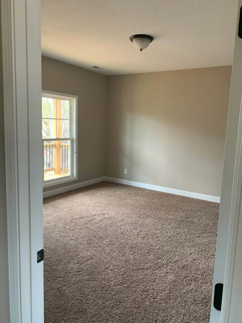 Neutral-toned carpeted room with white walls, ceiling-mounted modern light fixture, large window with white trim and railing, simple baseboard molding