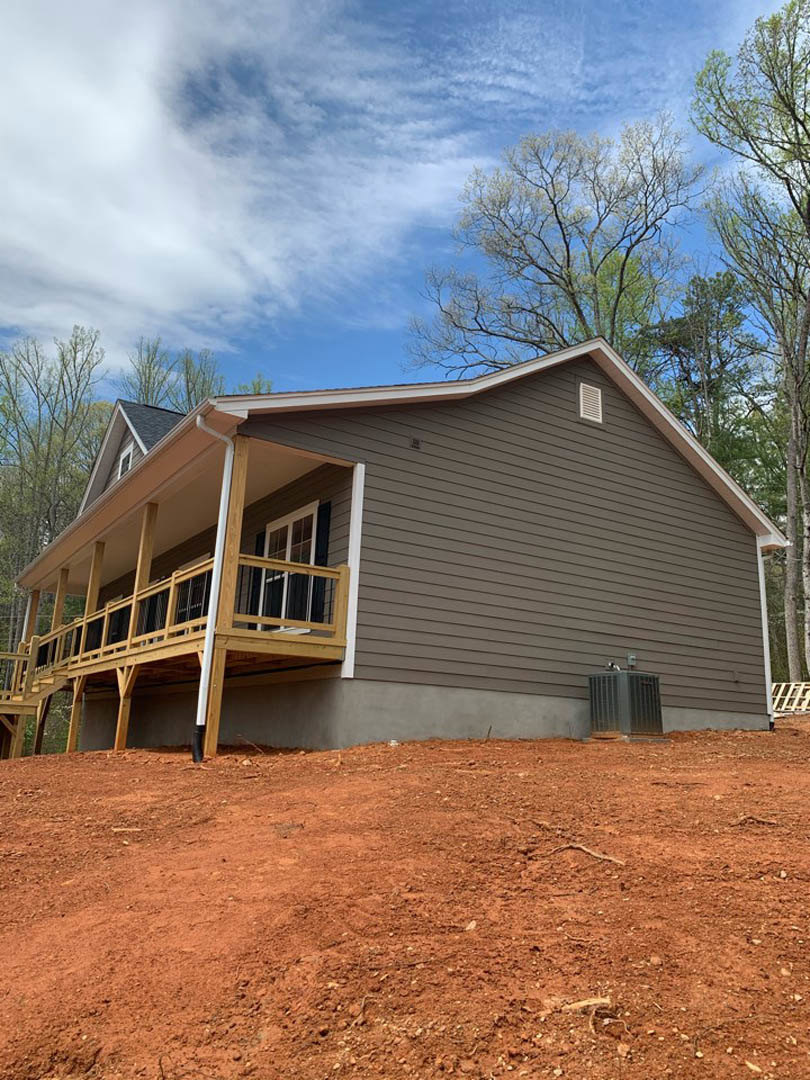 White siding house with covered front porch, wooden deck, metal vent, dirt landscaping, and tree under blue sky