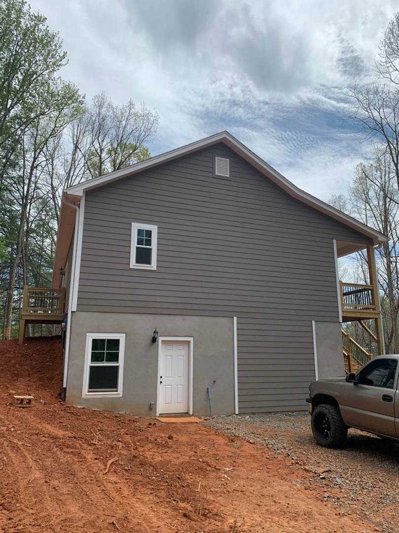 Grey siding house with attached garage, white front door, white-framed windows, parked pickup truck in driveway, cloudy sky overhead, trees in background