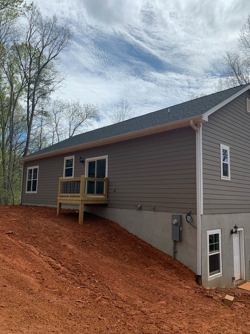 Two-story house with light siding, large windows, and a wooden deck overlooking a sloped dirt hill; white door with panel and railing visible; trees and cloudy sky in background.