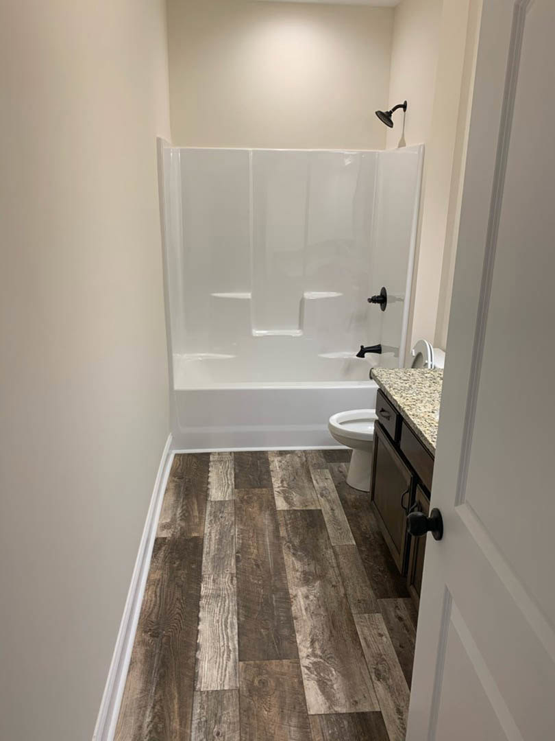 Bathroom with wood flooring, glass-enclosed shower featuring black shower head, white sink, and toilet; neutral walls and modern fixtures.