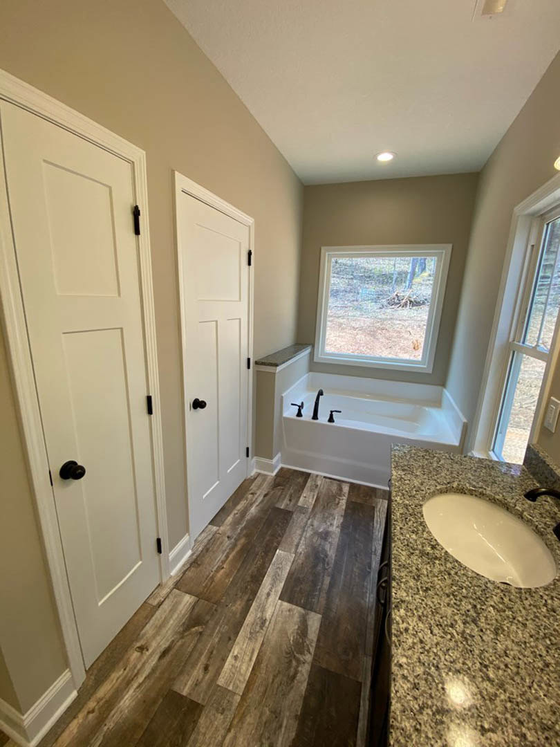 Marble bathtub beneath large window overlooking forest, white oval sink set on granite countertop, wood flooring, white doors with black handles, tile walls