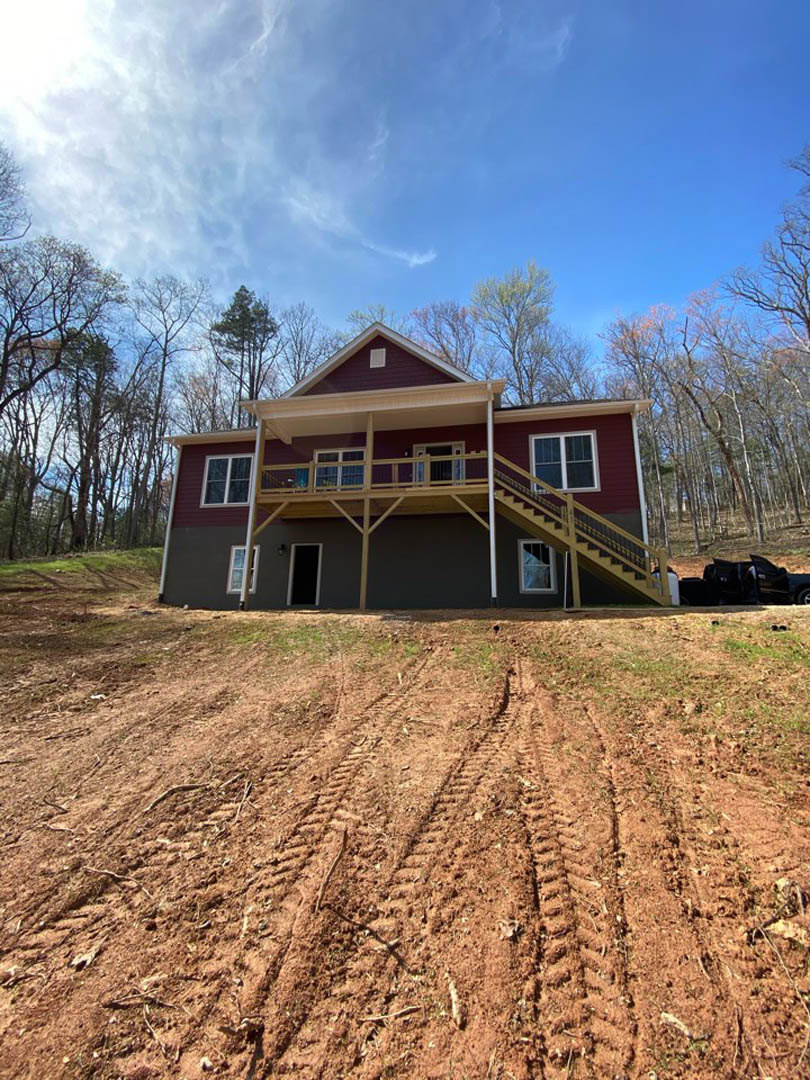 Two-story house with white-framed windows, covered porch, and balcony, situated on a dirt hill with tire tracks; black car parked nearby under a partly cloudy blue sky.