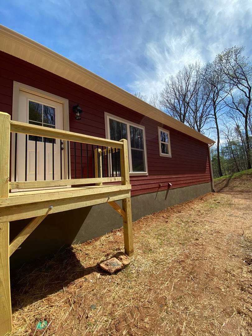 Red house with wooden porch and railing, fenced yard, grassy lawn, trees in background, cloudy sky