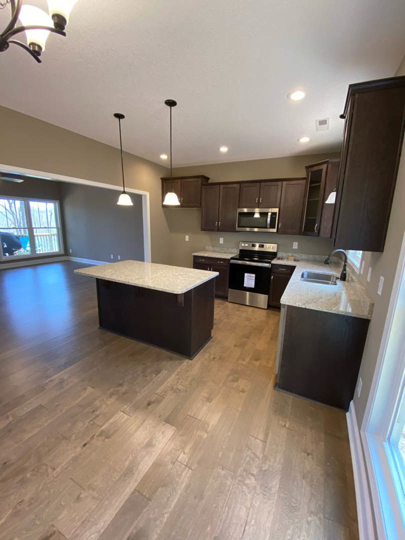 Kitchen with dark wood cabinets, wood laminate flooring, black and silver dishwasher, stone countertop, stainless steel sink, window with railing and person visible outside
