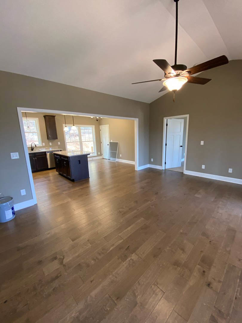 Open living room with hardwood floors, ceiling fan with light, white door with black handles, and adjacent kitchen featuring white cabinetry and countertops