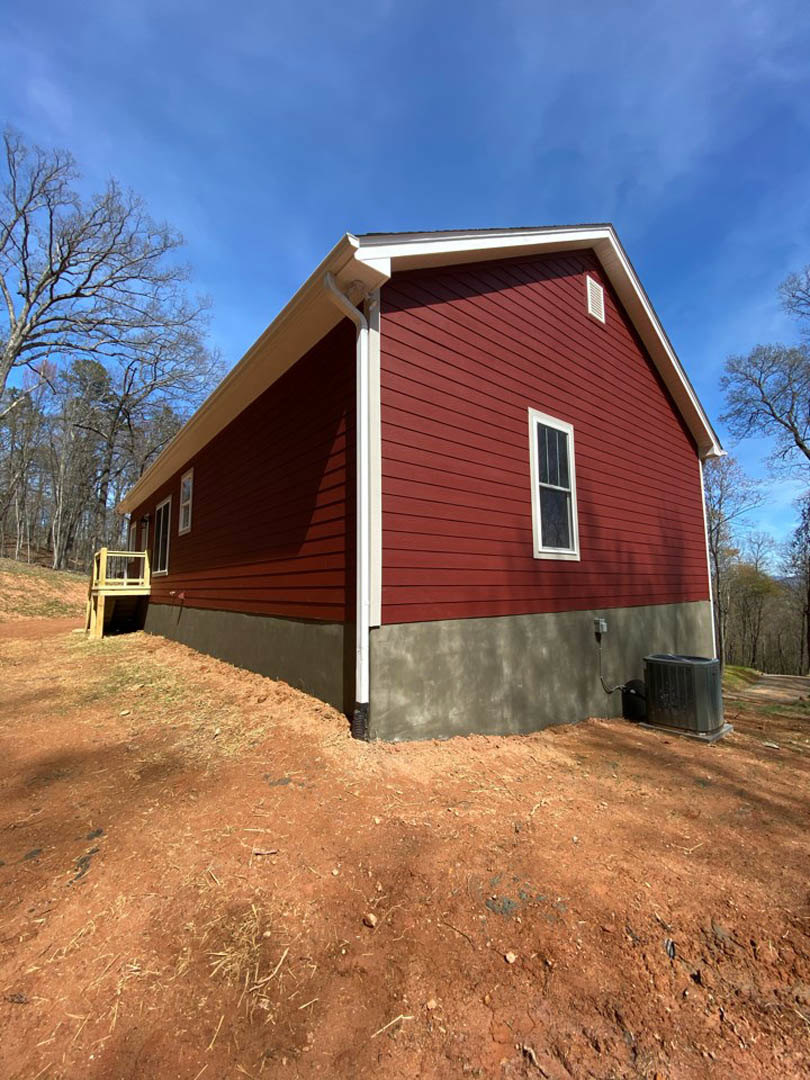 Red house with white roof and trim, wooden platform with railing, white-framed window, leafless tree, black box on ground, cloudy sky.
