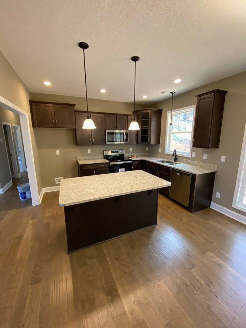 Spacious kitchen featuring a large central island with stone countertop, wood flooring, white cabinetry, stainless steel sink, and tile backsplash.