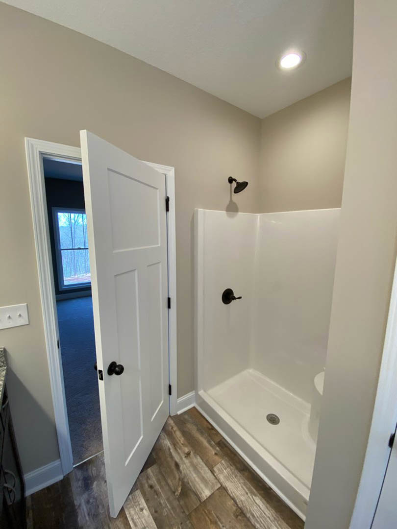 Modern bathroom featuring a white paneled door, tiled shower with floor drain, ceiling light, white wall switch with knobs, and window showing blue sky.