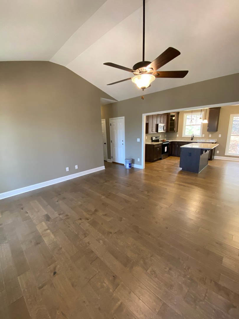 Open concept living room with hardwood floors, ceiling fan with light, white framed window, white door with black handle, and adjacent kitchen featuring a close-up of a countertop