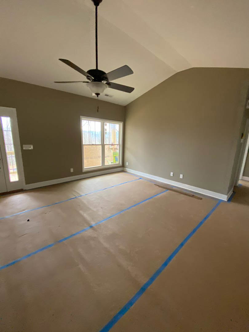 Room under construction with blue painter’s tape outlining the ceiling and floor, white-framed window letting in natural light, ceiling fan with integrated light fixture, plaster