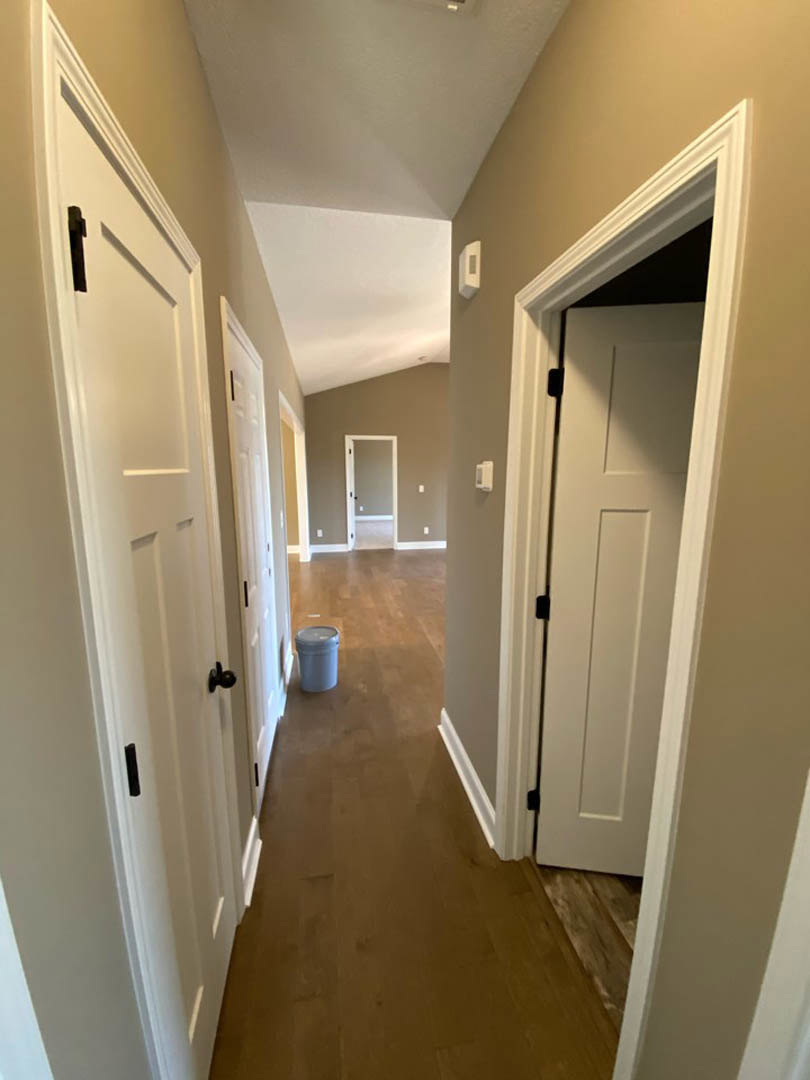 Hallway with wood flooring, white paneled doors featuring black handles, white walls with molding, and a grey trash can near the doorway