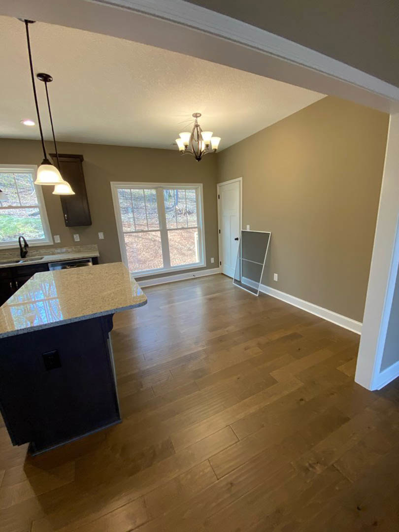 Open kitchen and dining area with rich hardwood floors, white cabinetry, light-colored countertops, five-light chandelier, white door with matching handle, and large window letting