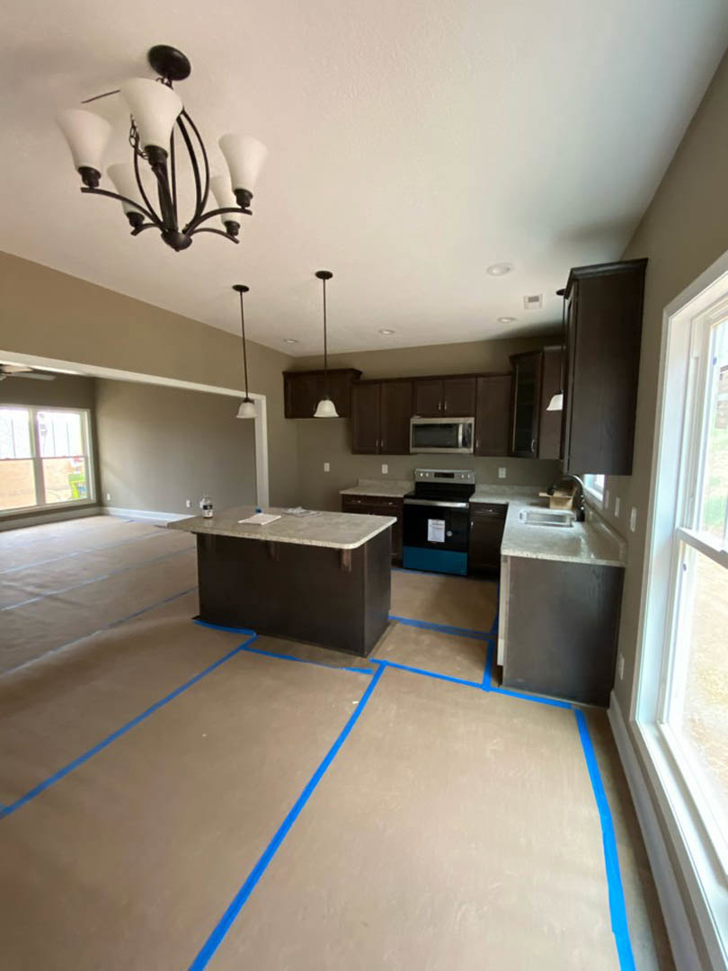 Modern kitchen under construction with blue painter’s tape marking edges on white cabinetry, light countertops, and hardwood flooring; black and blue oven, chandelier, and blue