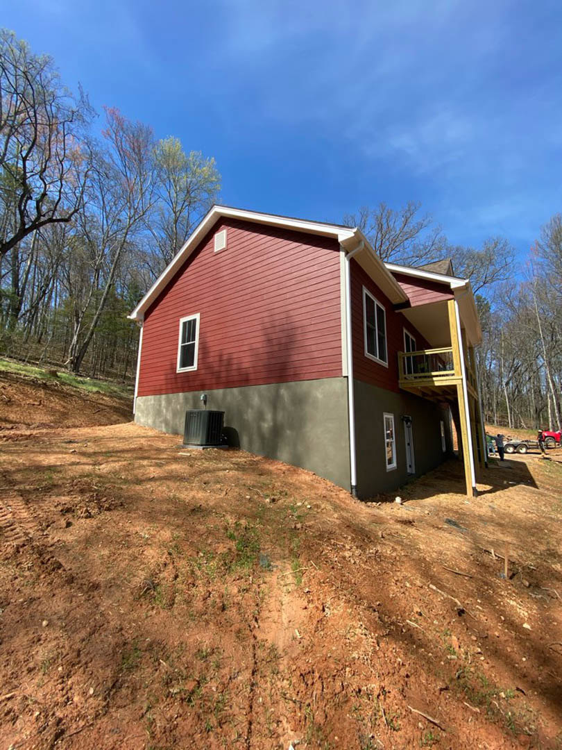Partially built house with exposed framing and red siding, elevated on a hill with a wooden deck, leafless tree nearby, red fence, black trash can, and scattered construction