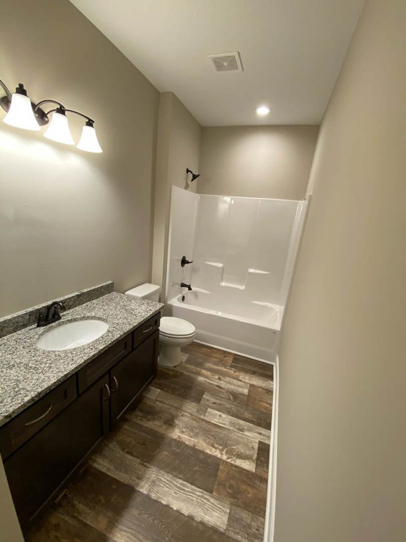 Bathroom with marble countertop vanity, chrome faucet, white bathtub, and toilet; three-light fixture above mirror; light-colored tile floor and walls.
