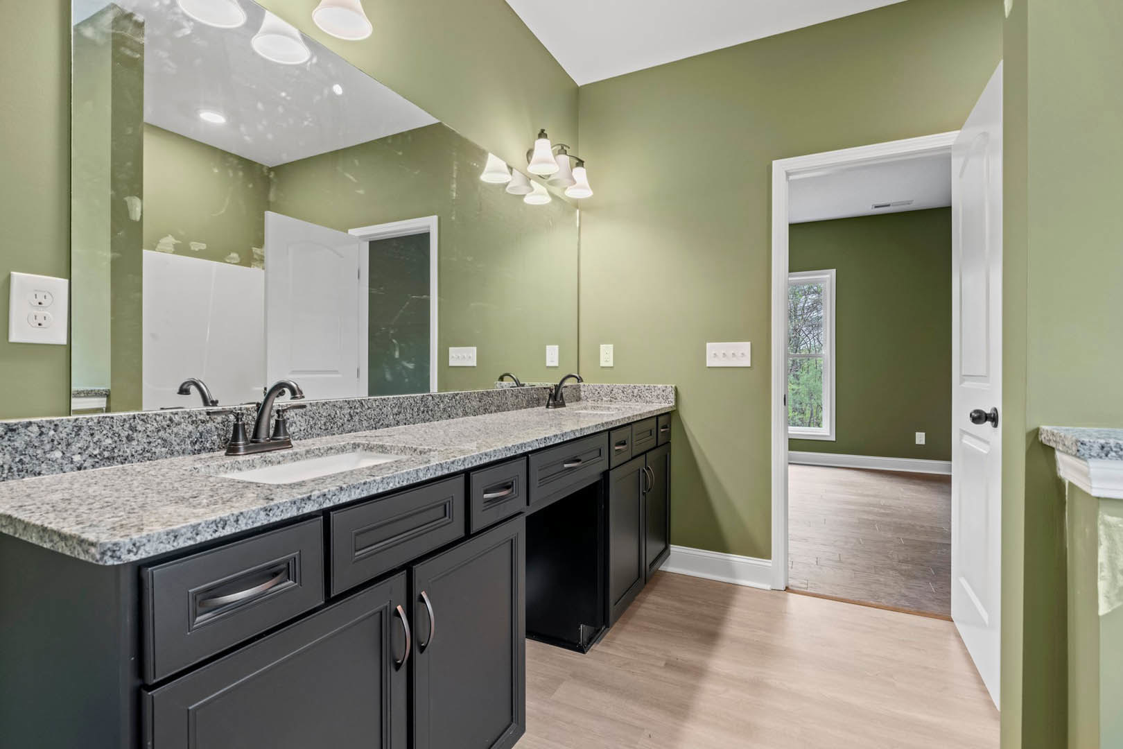 Bathroom with a wide mirror above a rectangular sink, light wood flooring, white door open to a green accent wall, modern outlet and light fixture, tile backsplash, and sleek