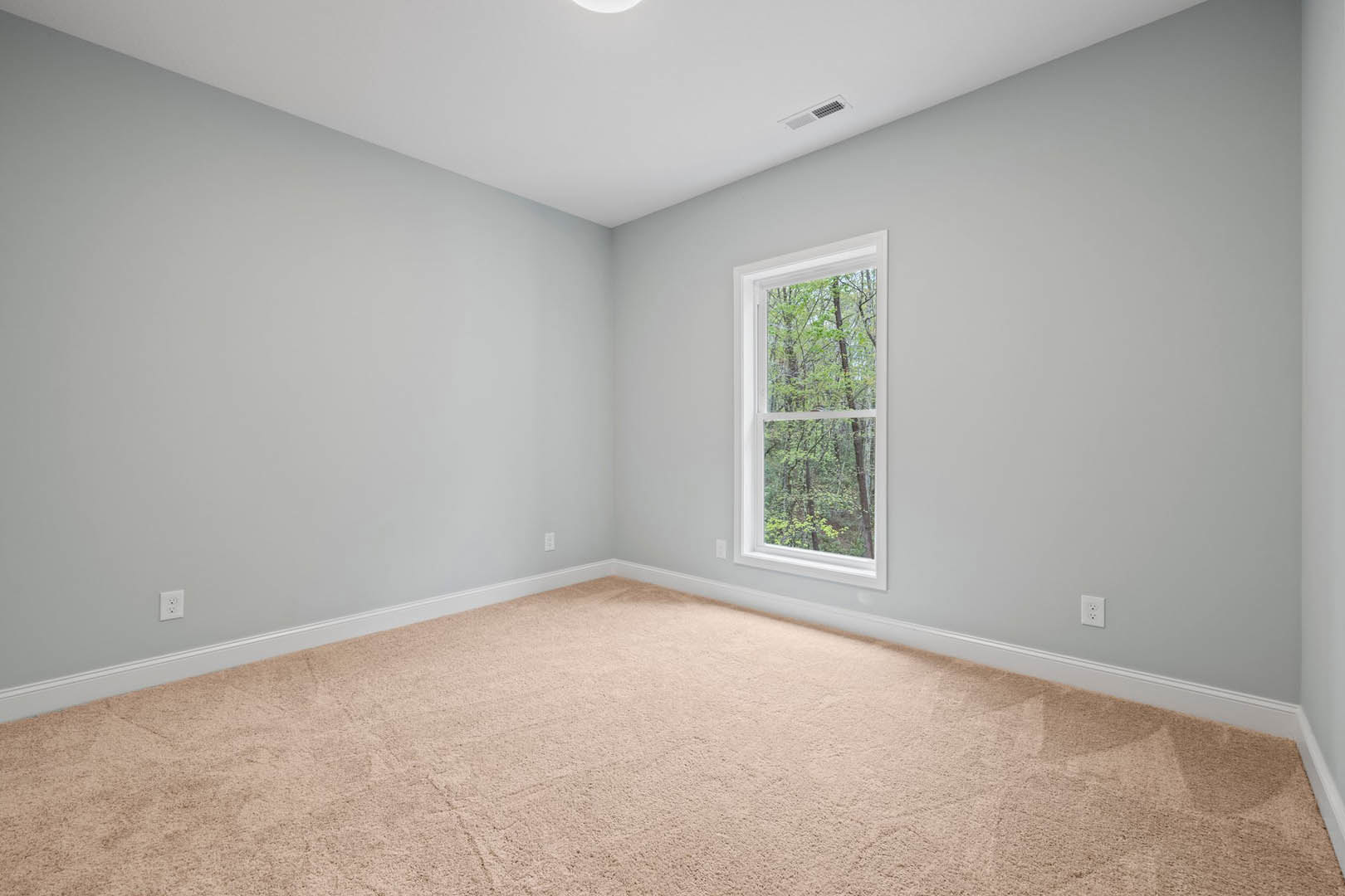 Carpeted room with large window overlooking leafy trees, white walls, ceiling vent, and crown molding