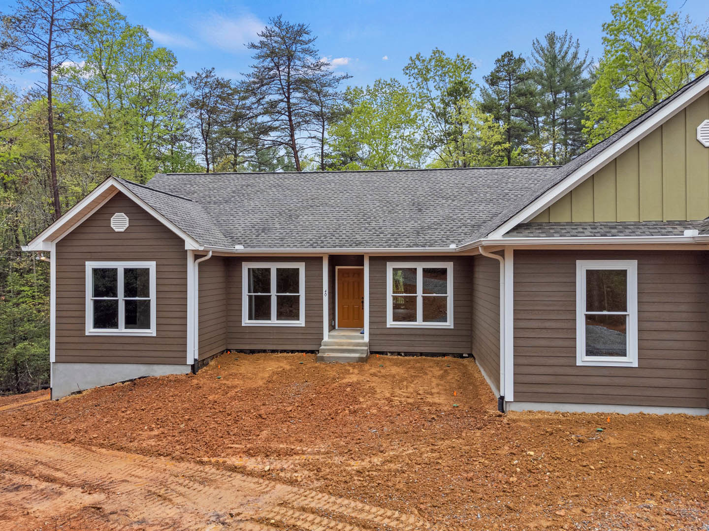 Brown roof and white trim house with large windows, white frames, and visible dirt landscaping surrounded by trees in the background