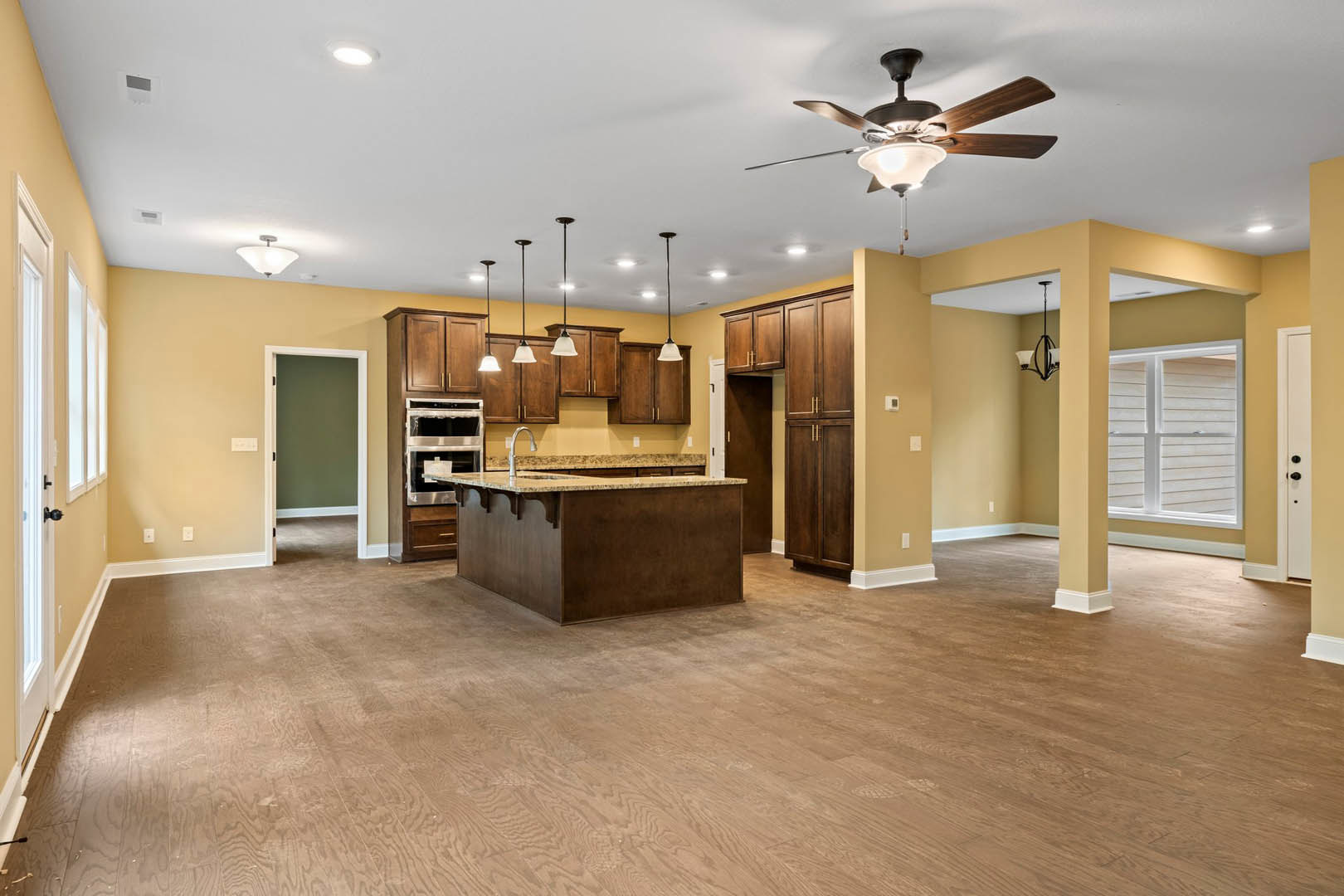 Open-concept kitchen and living room featuring a ceiling fan with light fixture, granite kitchen island, wood cabinetry, large window, and neutral flooring