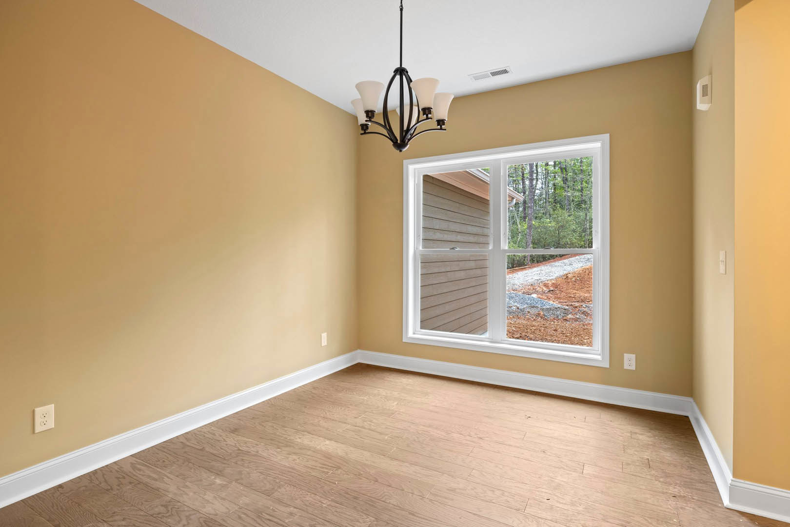 Sunlit room featuring a large window overlooking a dirt road and trees, wood flooring with white trim, white plaster walls, modern chandelier with white shades, and a white