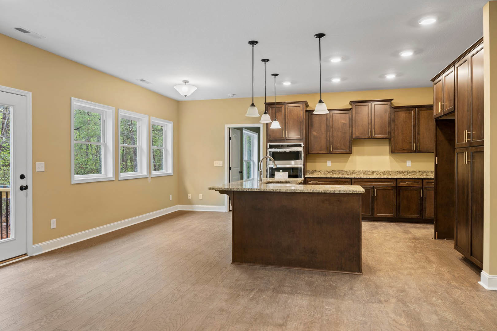 Open kitchen featuring a spacious island with quartz countertop, white cabinetry, hardwood flooring, row of windows overlooking trees, stainless steel faucet, and door with black