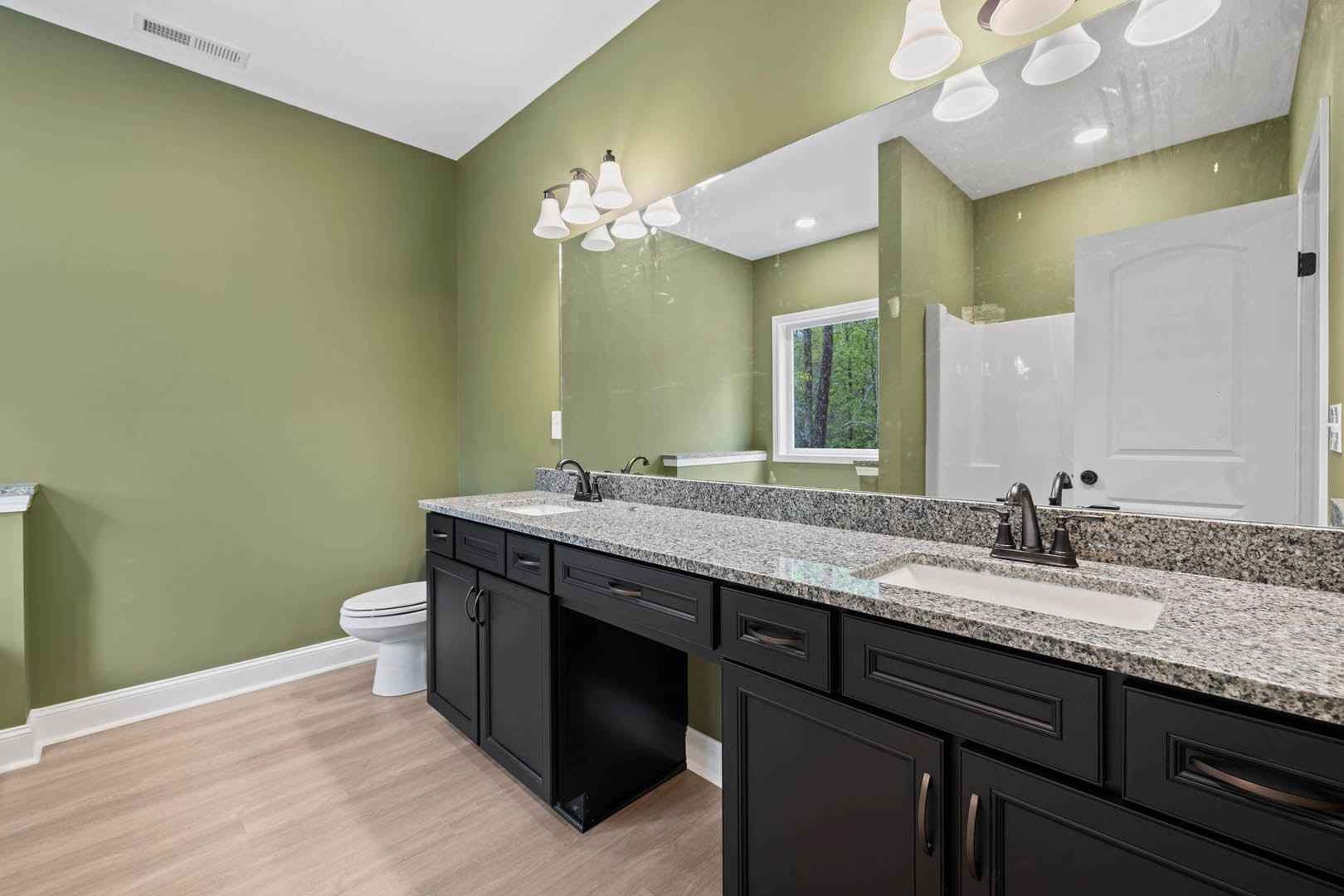 Bathroom with a wide mirror above a rectangular sink, white cabinetry with black hinges, chrome faucet, stone countertop, toilet, and window overlooking trees.
