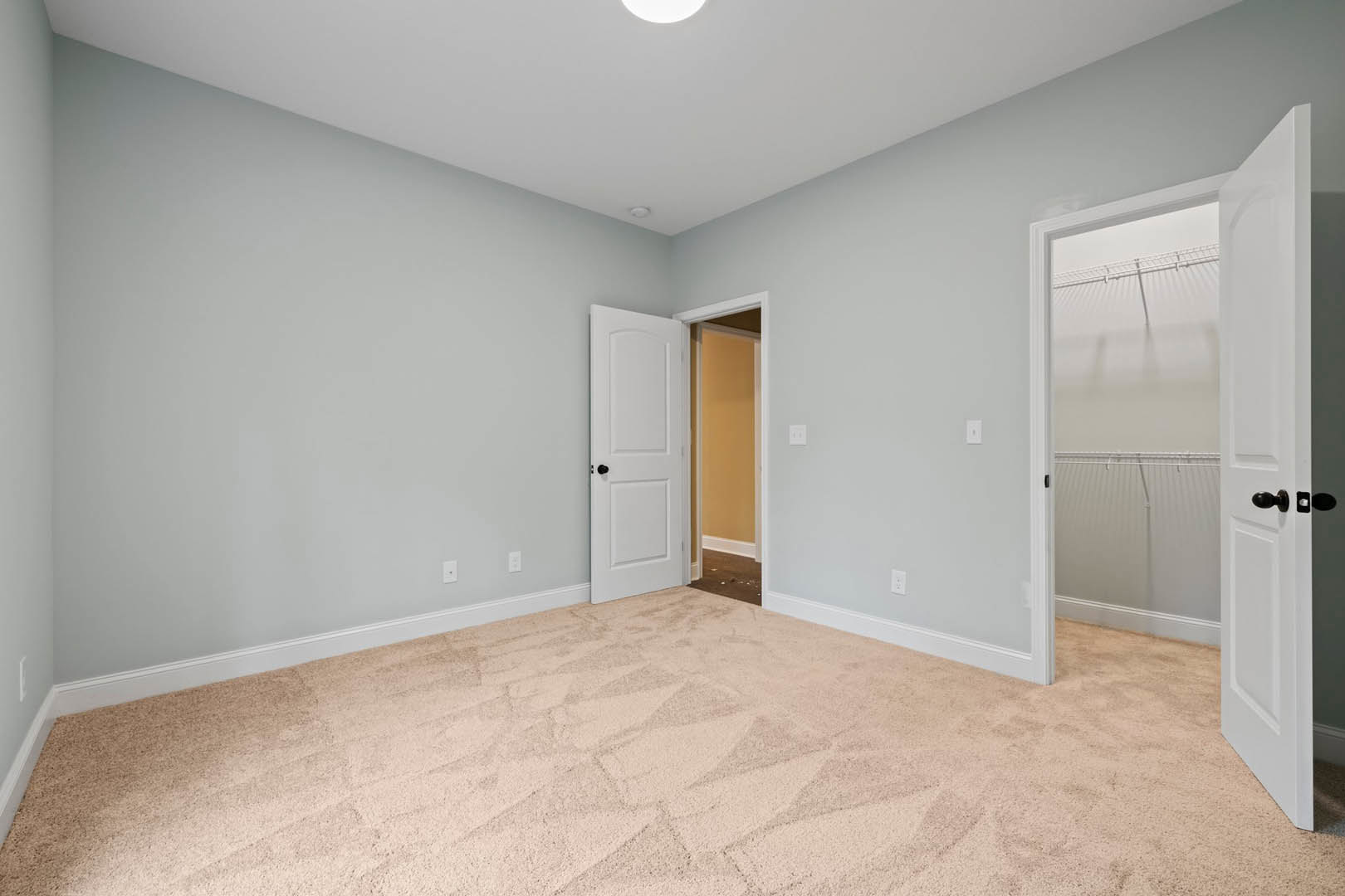 Patterned carpeted floor, white door with black knob open to adjoining room, white ceiling with round flush light fixture, close-up of window and door trim