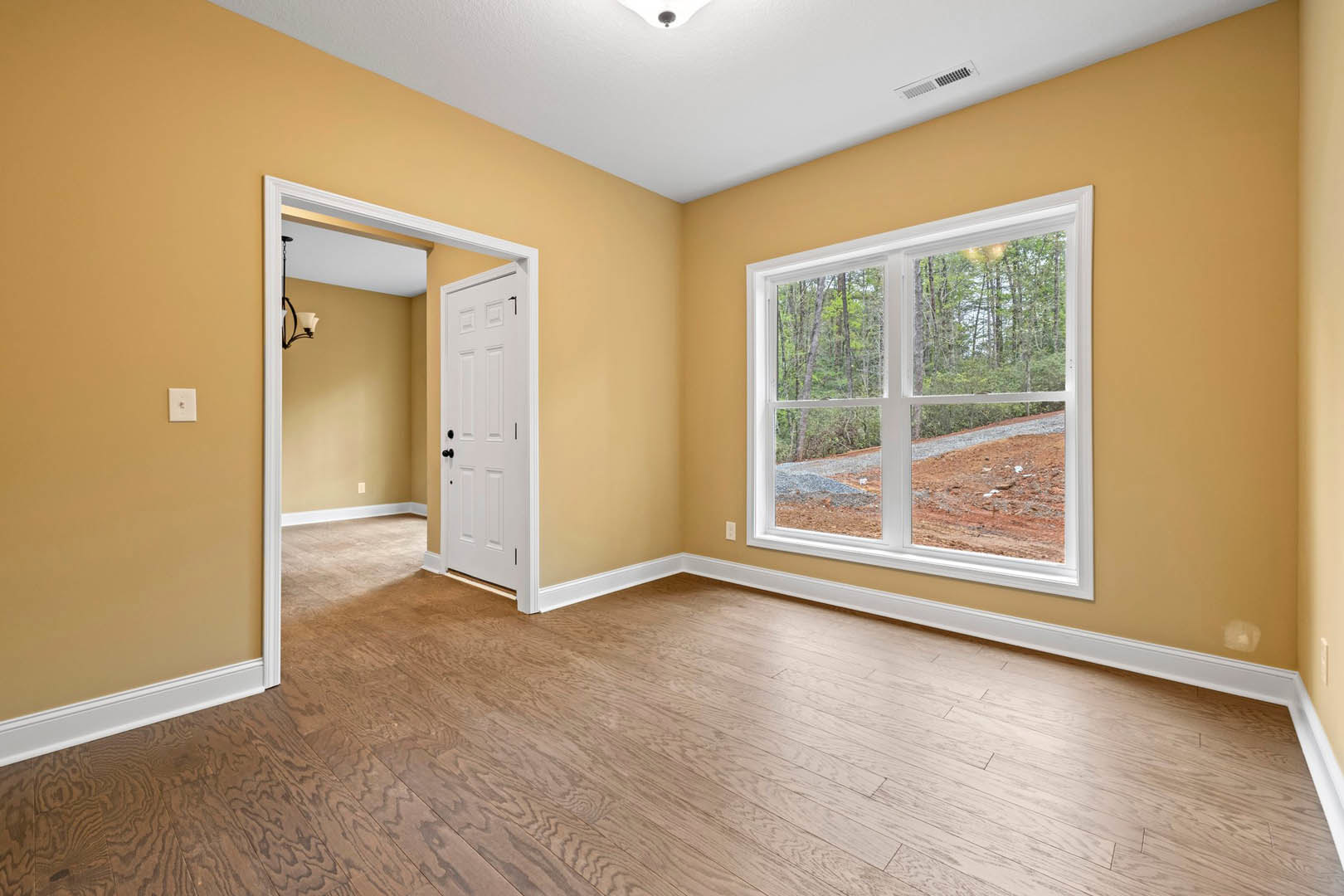Hardwood floor room with white door featuring black knobs, large window overlooking dirt hill and trees, white walls, ceiling vent, and close-up lamp.