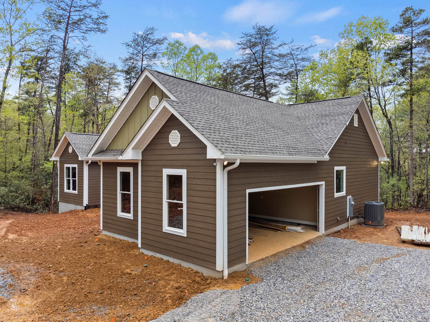 Partially built house with exposed framing, attached garage, white window frames, broken glass pane, stack of white planks on ground, surrounded by trees