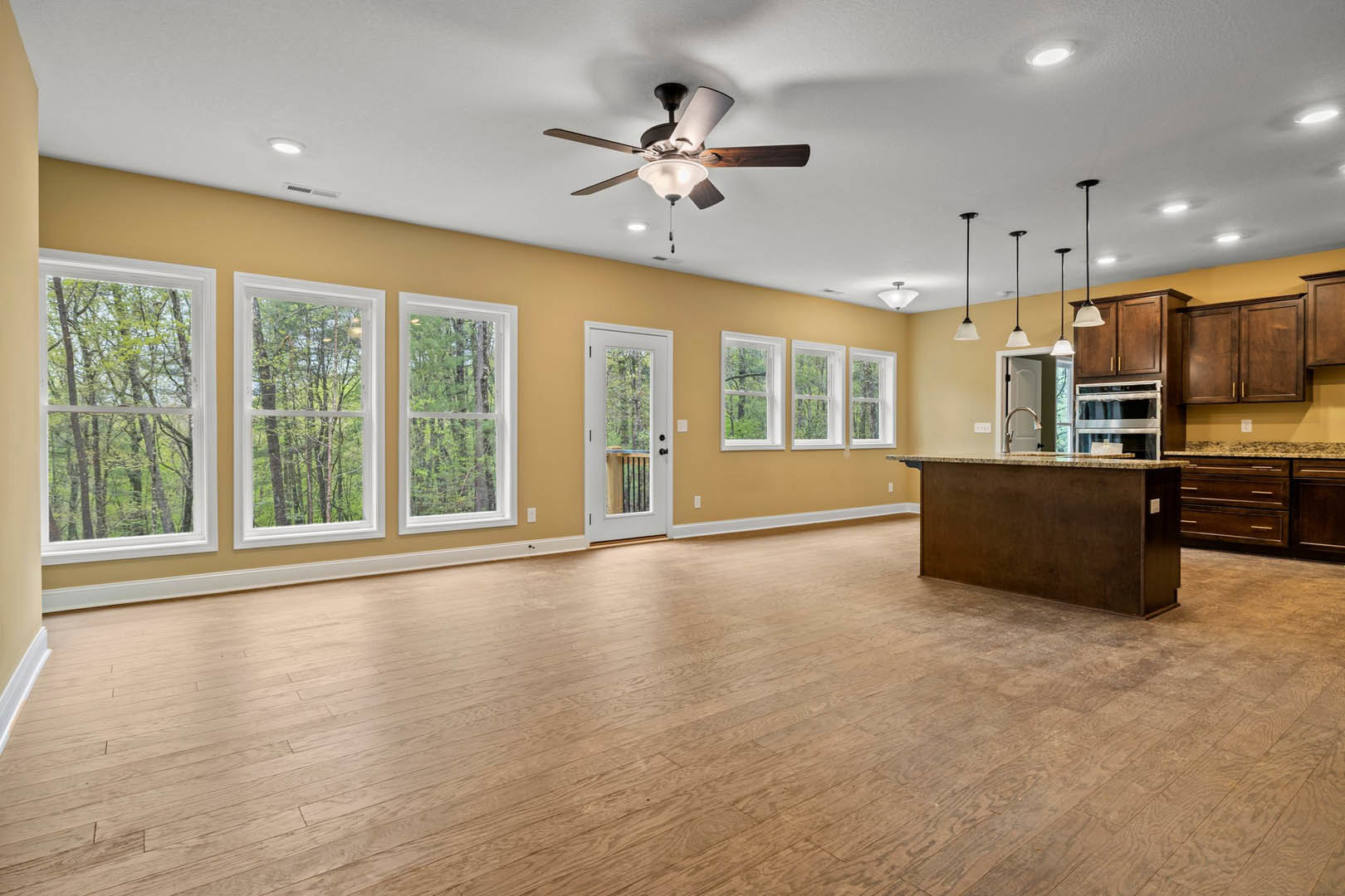 Open-concept kitchen and dining area with hardwood floors, white cabinetry, stainless steel appliances, ceiling fan with light fixture, large windows showing trees outside, and