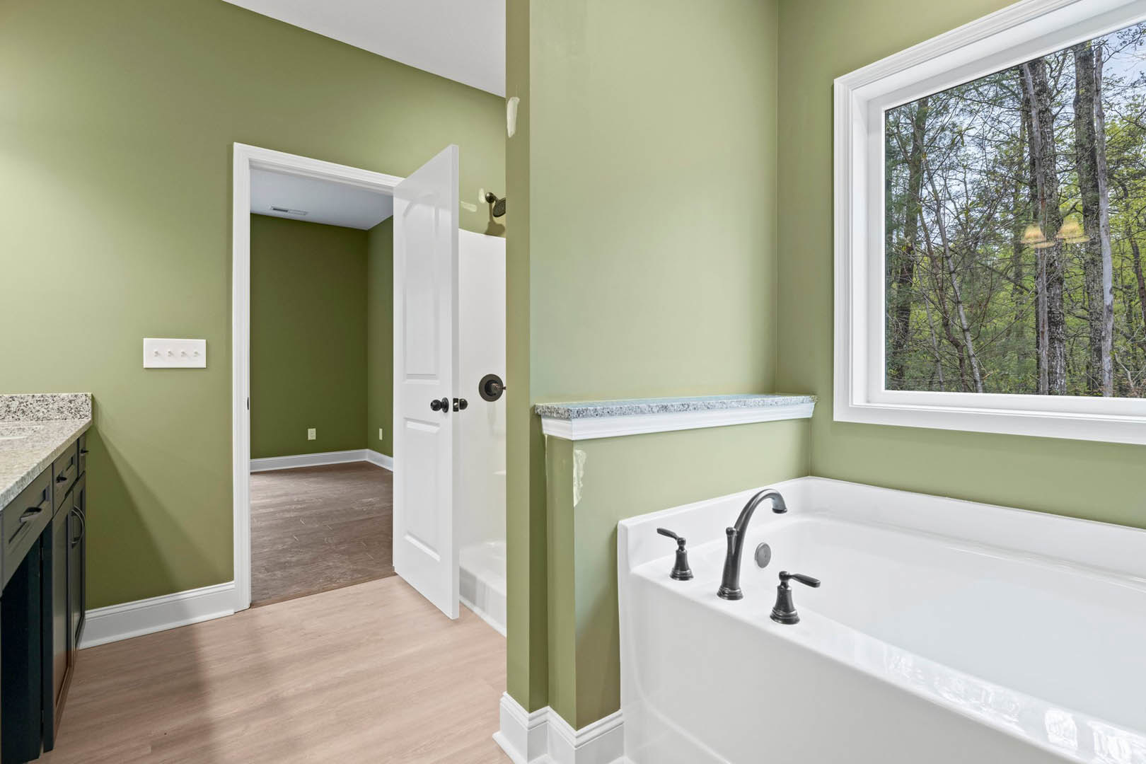 Freestanding bathtub with chrome faucet against a green accent wall, large window overlooking trees, wood flooring, white door, and white trim