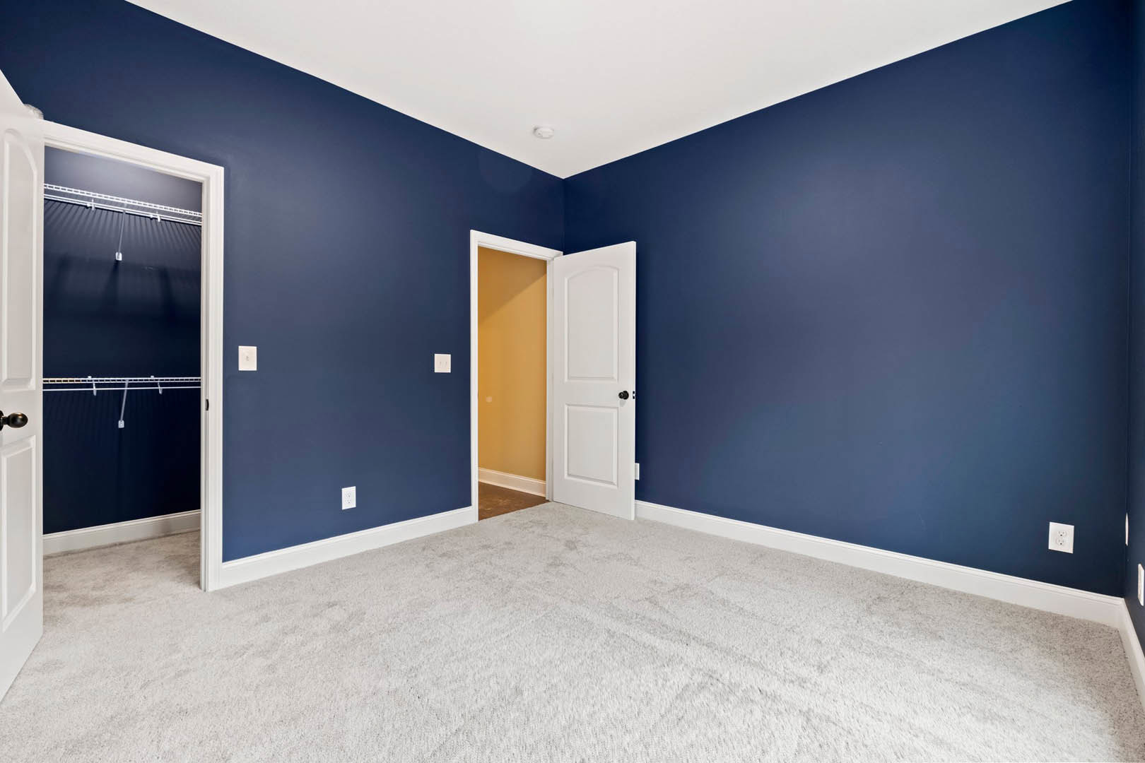 Blue walls and white carpet in a room with a white door featuring a black knob, white ceiling with smoke detector, yellow accent wall with white trim, and partial view of roof