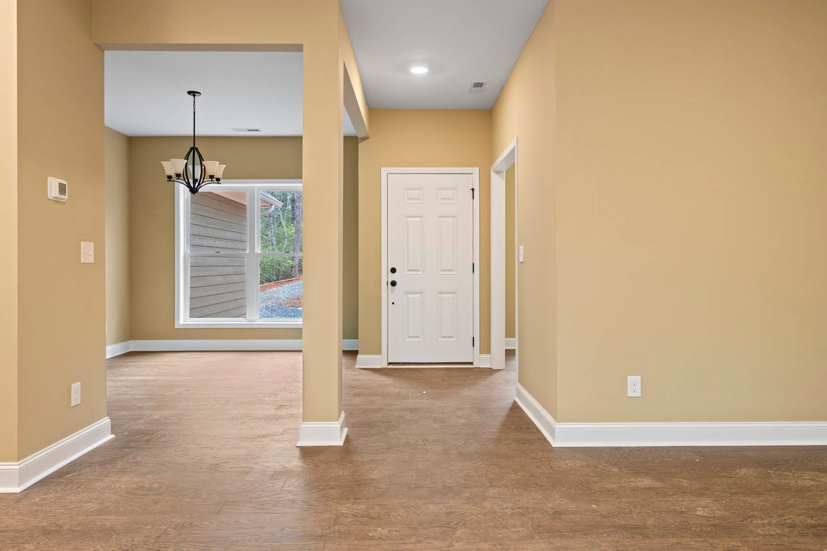 Hallway with light wood flooring, white walls, white door with black hardware, window offering outdoor view, white smart device mounted on wall, close-up of modern chandelier