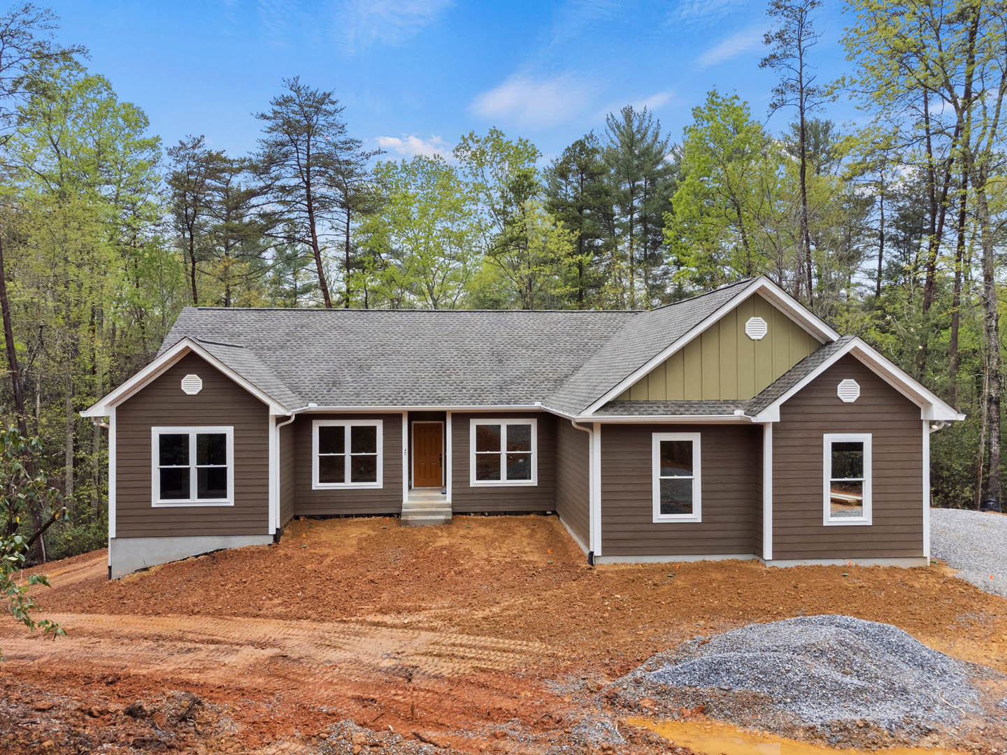 Partially built wood-frame house with brown roof, white-trimmed windows, and unfinished door, surrounded by tall trees and dirt patch in front