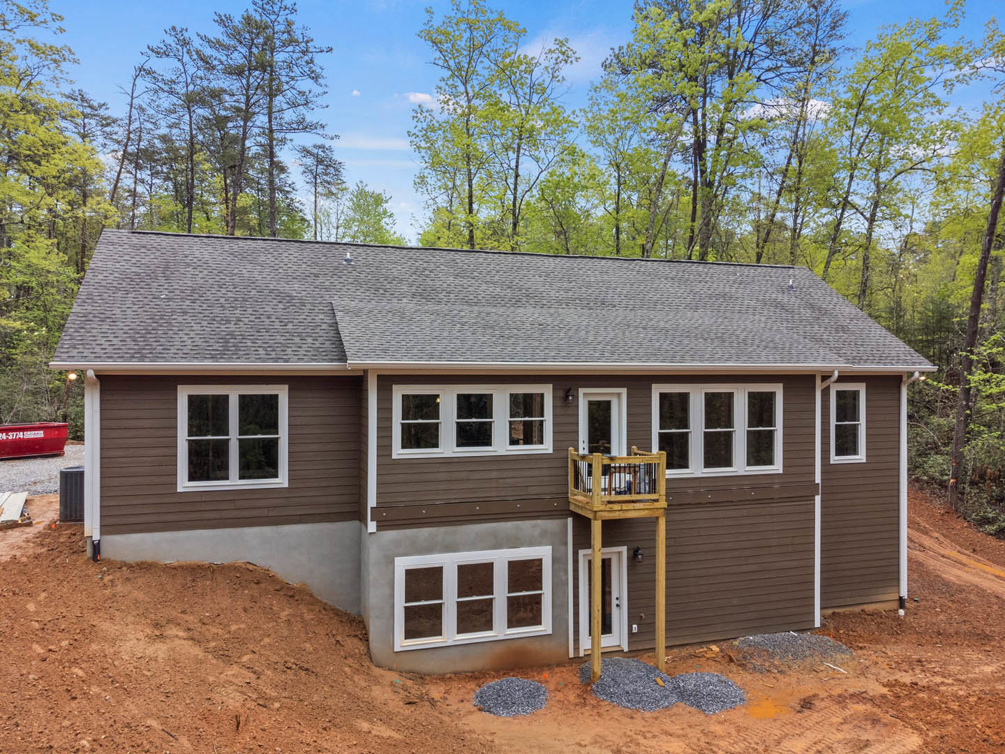 Wood-framed house under construction with exposed balcony, white door, close-up window, pitched roof, and surrounding trees; red dumpster with white text in foreground
