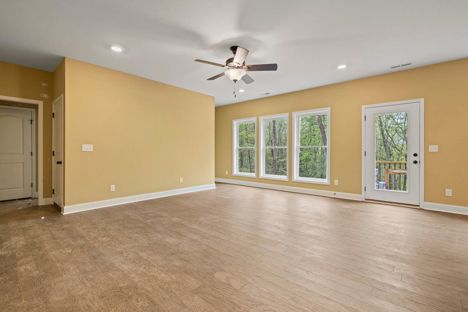 Ceiling fan with light fixture, hardwood floor, white door with black handle, glass door opening to deck, windows showing trees outside, plaster walls and ceiling