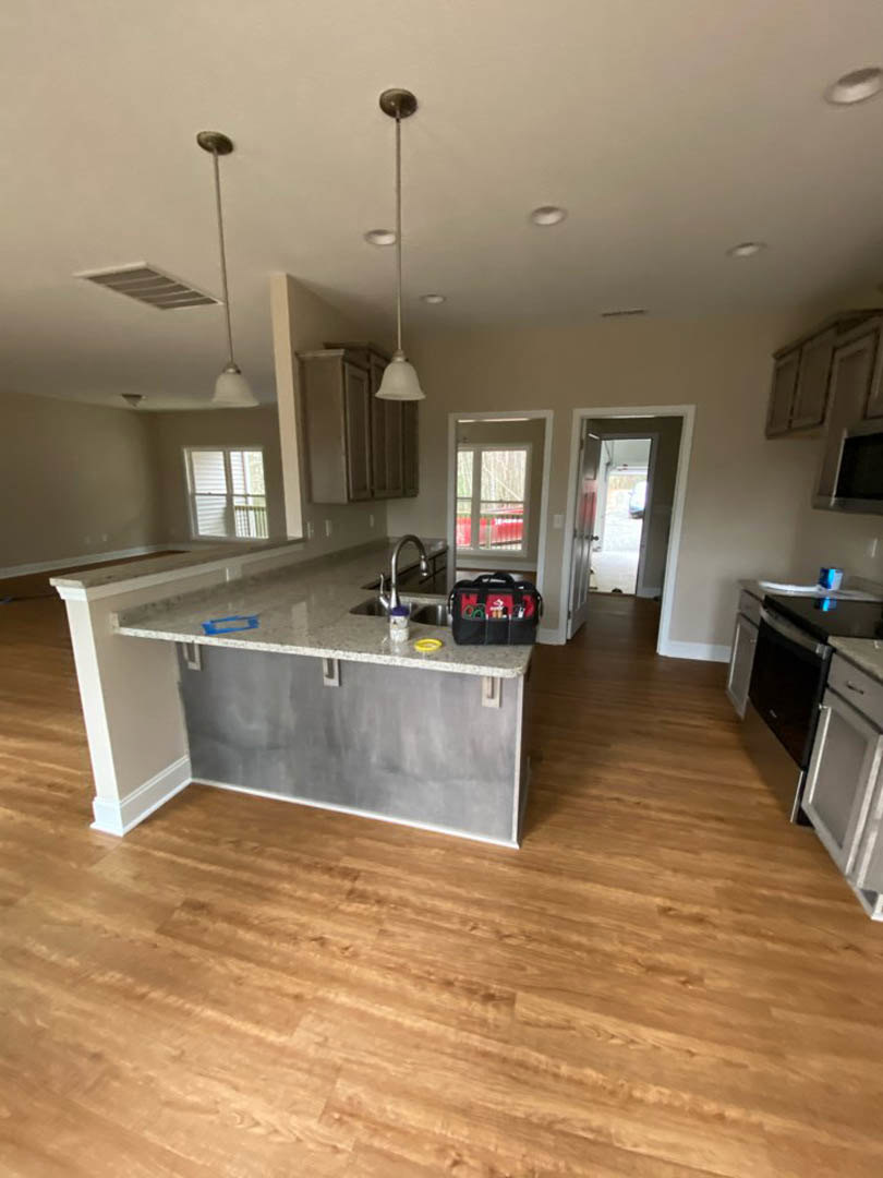 Kitchen with light wood flooring, white cabinetry, stainless steel sink and faucet, stone countertop, window with white blinds, black bag with red and white items, toolbox on