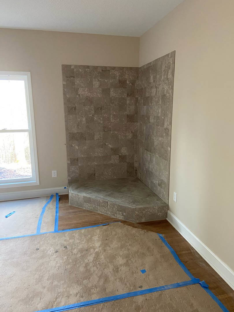 Corner of a room featuring a tiled shower stall with light-colored tile walls and floor, adjacent to a window and carpeted area
