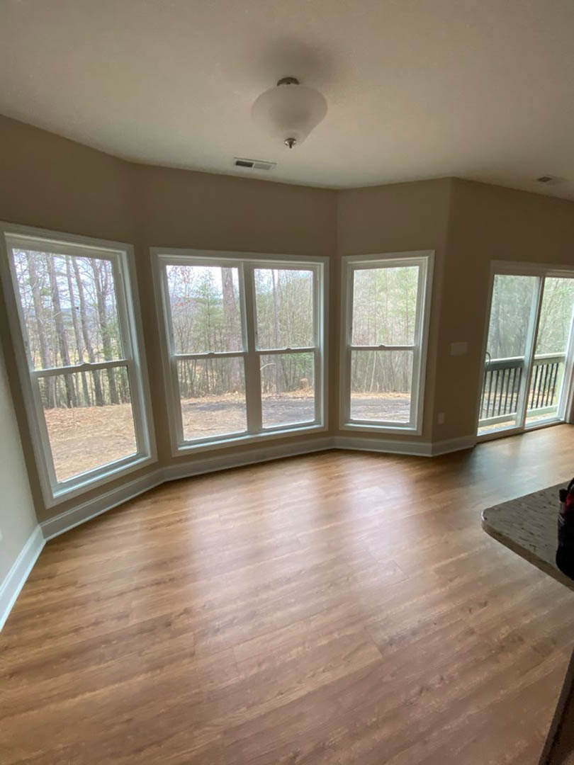 Hardwood floor room with large windows, white frames, ceiling light fixture, glass door opening to outdoor railing, trees visible outside