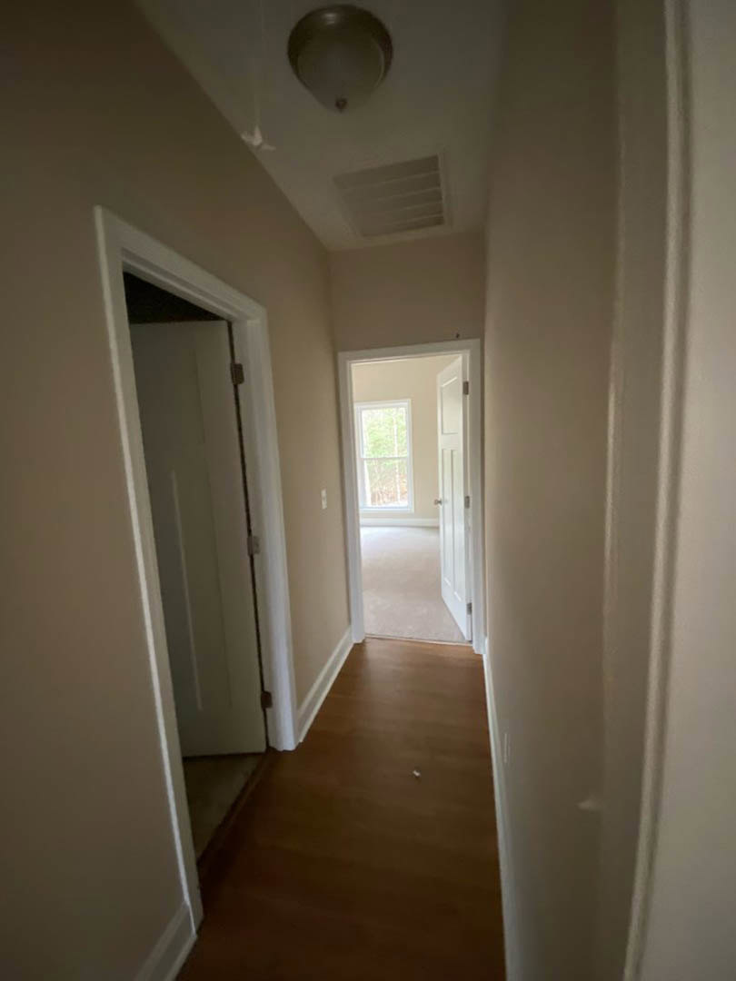 Hallway with light wood flooring, white walls, and an open white door; white ceiling with vent and crown molding visible