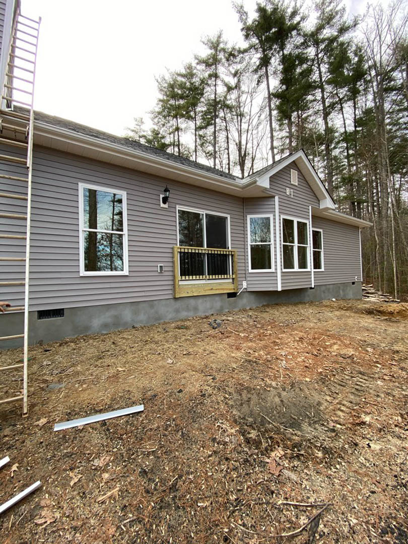 Grey siding exterior with white-framed window, white ladder leaning against wall, dirt and debris scattered on ground, trees visible through window in background