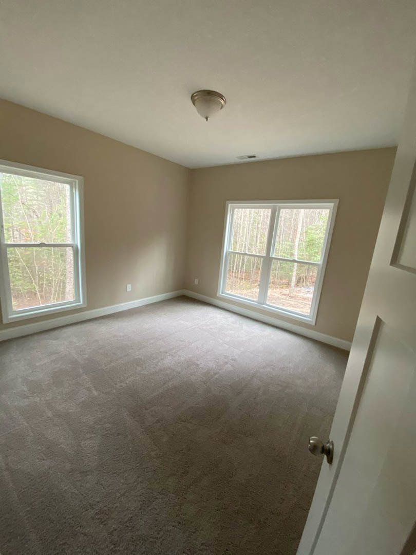 Carpeted room with white walls, large windows showing trees outside, white door with silver knob, and modern ceiling light fixture
