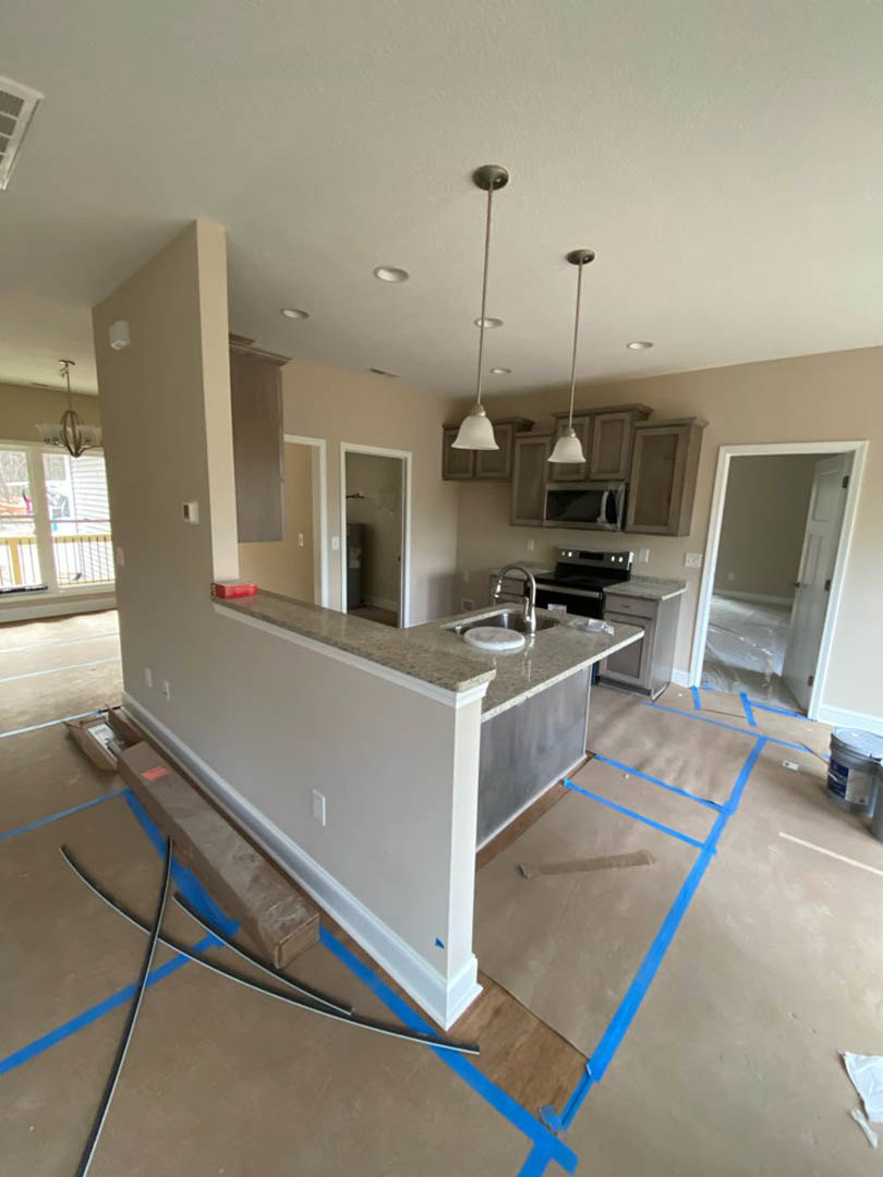 Modern kitchen under construction with white walls, unfinished cardboard-covered floor secured by blue tape, stainless steel sink set in a light countertop, door wrapped in