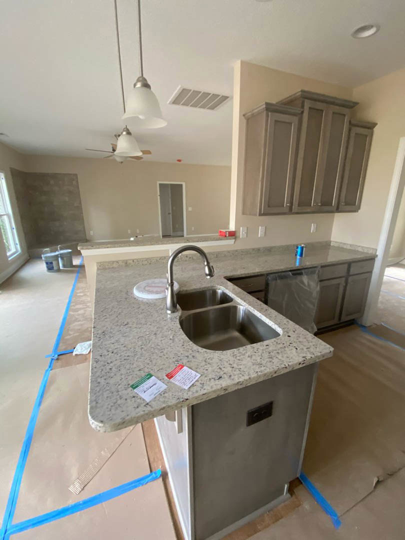 Modern kitchen featuring a central island with a stainless steel sink and faucet, white cabinetry along the wall, tile backsplash, black electrical outlet, and a white door with a