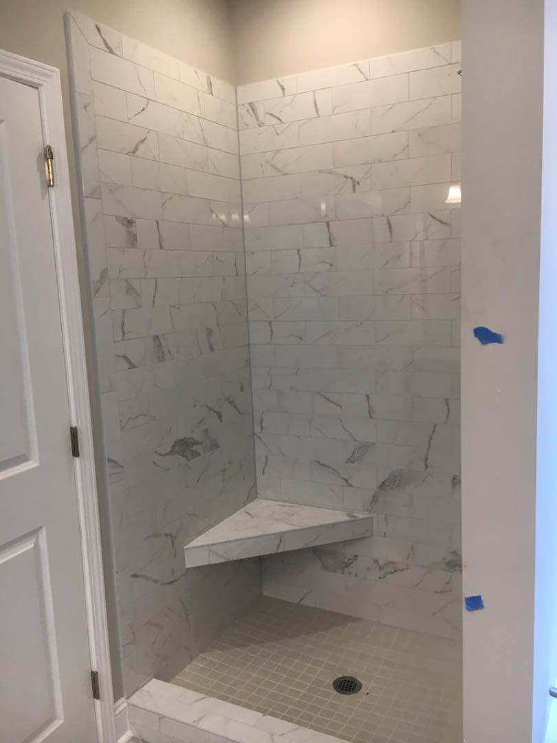 White tile shower featuring a built-in bench, corner shelf, and linear drain, adjacent to a white countertop and wall with a vertical white pole