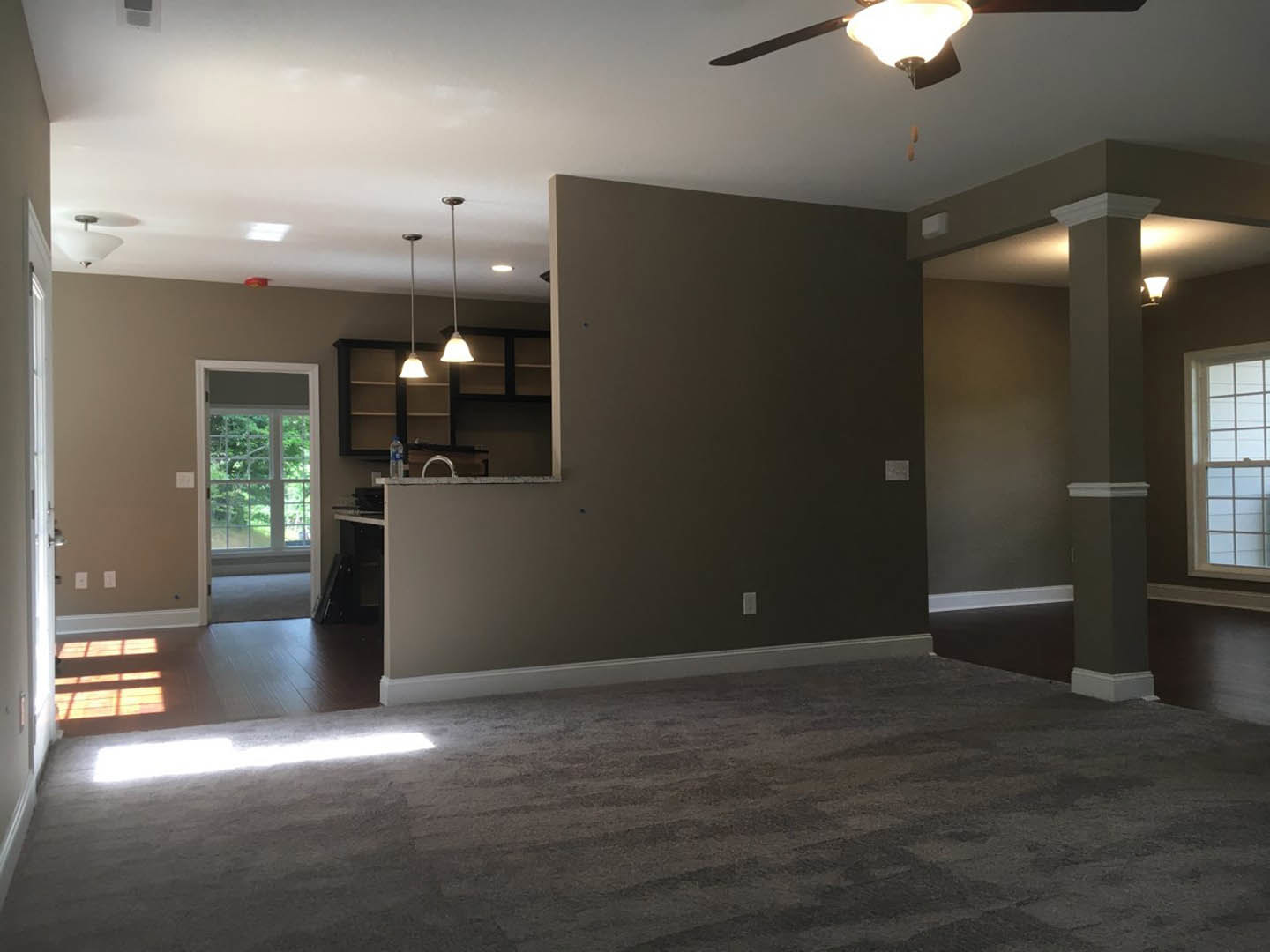 Open-concept living room and kitchen with carpeted floor, multi-pane window overlooking trees, ceiling light fixture, and neutral walls