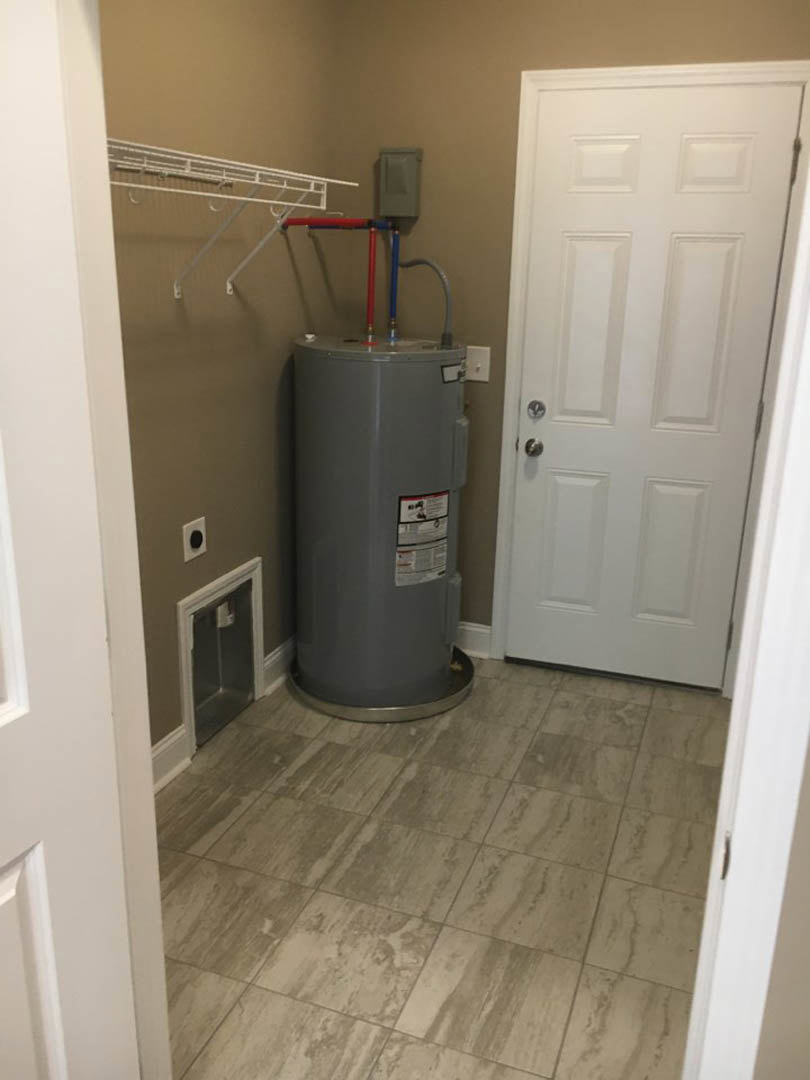Grey cylindrical water heater beside a white door with silver handle, tile flooring, small trash can, wall-mounted white switch, and water dispenser in a utility room.